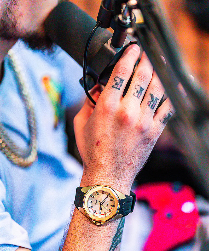 Hand with tattoos wearing a gold watch and gripping a microphone.