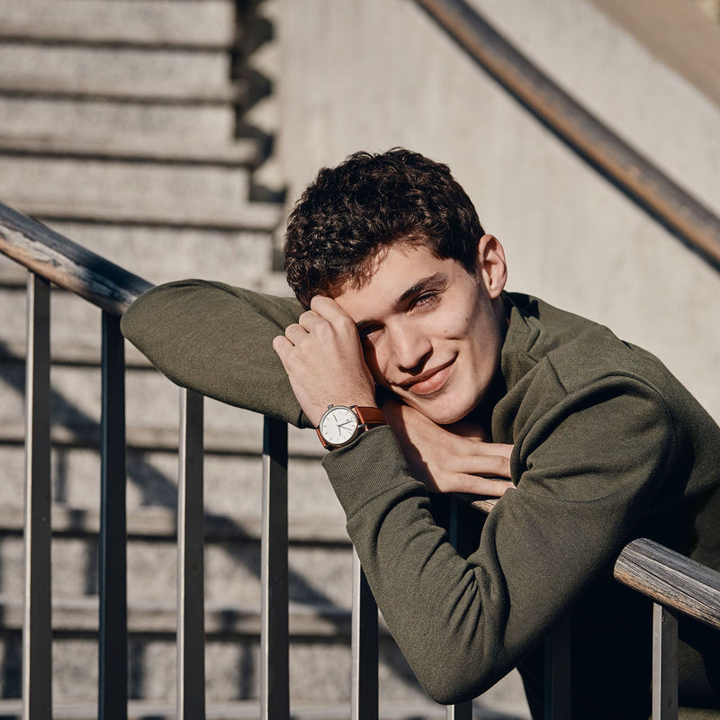 Young man with curly dark hair wearing an olive green sweater and a wristwatch, leaning on a railing.